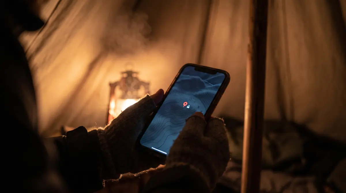 Gloved hands hold a phone inside a wall tent at night. A lantern glows behind it, and the screen shows a dark topographic map with one red marker.